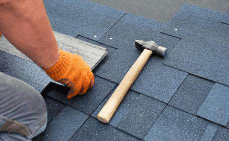 Roofer installing asphalt shingles with hammer and orange gloves on residential roof