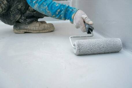 Worker in gloves applying gray paint with roller on white surface floor coating