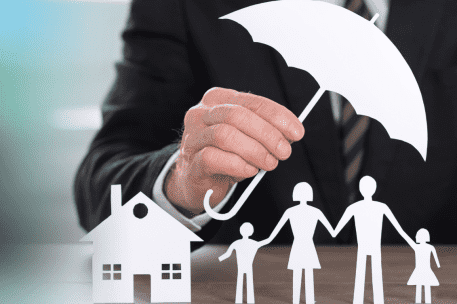 Hand holding white umbrella over paper cutouts of house and family symbolizing insurance protection