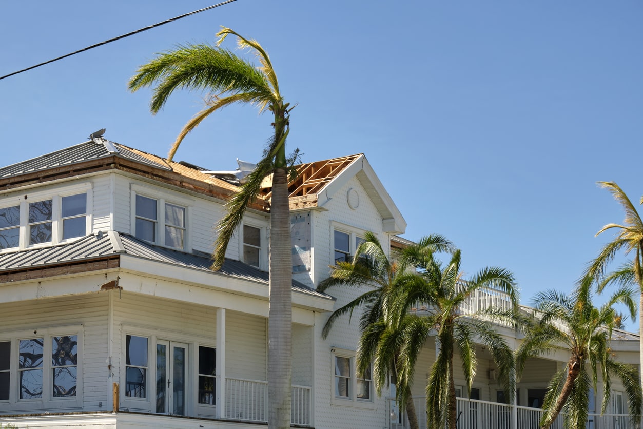 Hurricane damaged white house with torn roof and bent palm trees under blue sky